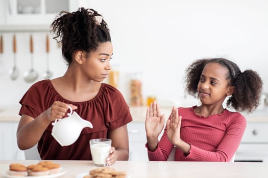 Lactose intolerance concept. Black teen girl refuse to drink milk, sitting at kitchen table with her mom, eating homemade pastry, african american woman mother pouring milk into glass