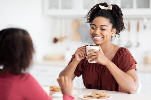 Pretty black woman drinking tea with her teen daughter, holding coffee mug, looking at kid and smiling, kitchen interior, copy space. Mother and daughter communication, motherhood concept