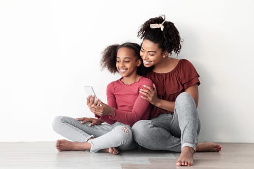 Beautiful black mother and daughter using mobile phone together, sitting on floor, hugging and taking selfie for social media, enjoying time together at home, white empty wall backgound, copy space