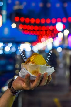 Hand holding coconut and mango ice cream in coconut shell against lights of Jalan Alor street food market in Kuala Lumpur