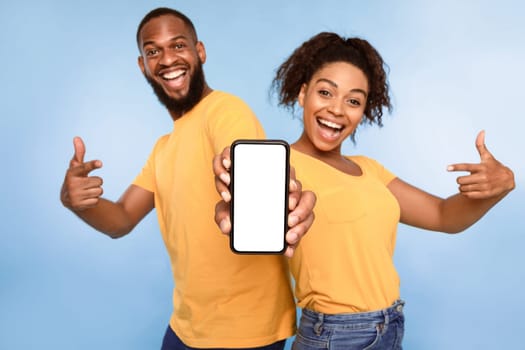 Mobile application ad. Excited African American couple pointing at cellphone with blank screen, recommending new app on blue studio background, selective focus. Mockup for website