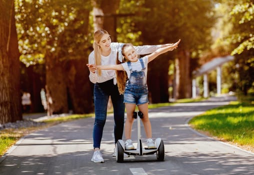 Mom Teaching Daughter To Ride Segway Having Fun In City Park Outdoors. Weekend Leisure Concept. Free Space