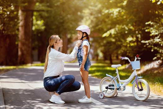 Mother Putting Helmet On Daughter Teaching Her To Ride A Bicycle In Park On Weekend. Free Space For Text