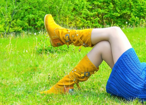Slim female legs in yellow leather boots on bright green grass with dandelions in the summer.
left leg rests on right, blue knitted dress