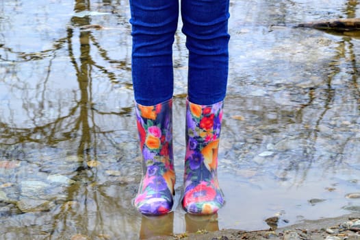 Women's legs in blue jeans and floral rubber boots in the puddle with reflection