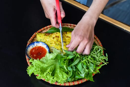 Hands with scissors cutting Vietnamese pancake Banh xeo with sea food on black background top view