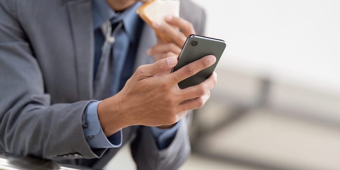 A close up view of a young Asian businessman using his smartphone while enjoying a snack, showcasing a blend of work and personal life in a sleek office setting.