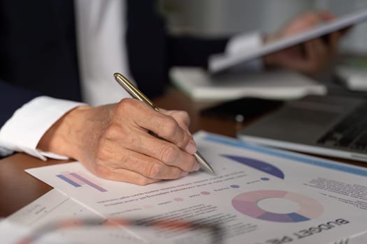 An elderly businessman meticulously reviewing budget reports and graphs, writing notes at his office workspace.