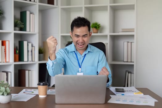 An excited Asian businessman celebrates a victory at his desk, raising his fists in joy while engaging with his laptop in a contemporary office setting.