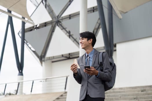 A young Asian businessman in a suit, smiling as he enjoys a snack and checks his smartphone, embodying a cheerful and balanced work life in a contemporary outdoor environment.