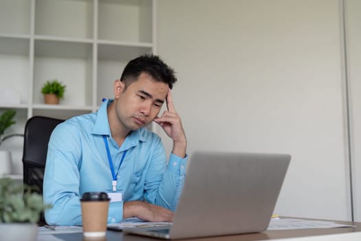 A thoughtful Asian businessman reflects at his desk, contemplating work challenges while staring at his laptop and holding a coffee cup in a contemporary workspace.