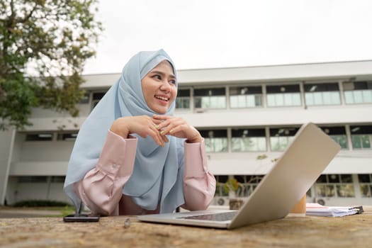 A delighted Muslim businesswoman in a hijab enjoying her work at a table, radiating positivity in an outdoor environment.