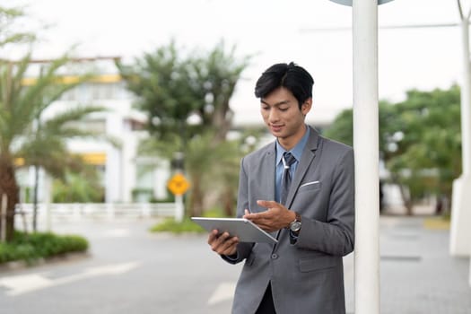 A young Asian business professional stands outdoors, smiling as he interacts with a tablet, highlighting engagement and the use of technology in a work environment.