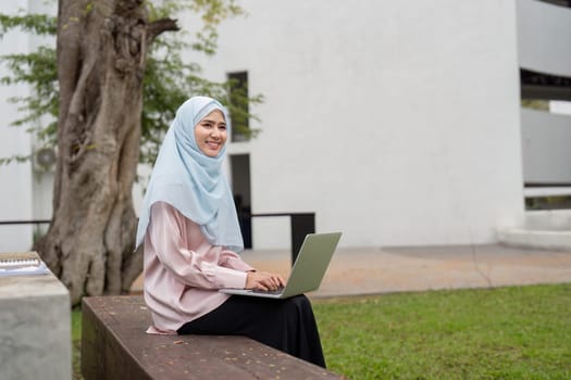 A cheerful Muslim businesswoman in a hijab, happily working on her laptop while seated on a bench in a green outdoor space.