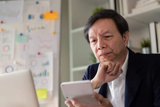 An elderly Asian businessman thoughtfully reviewing data on his smartphone in a contemporary office setting.