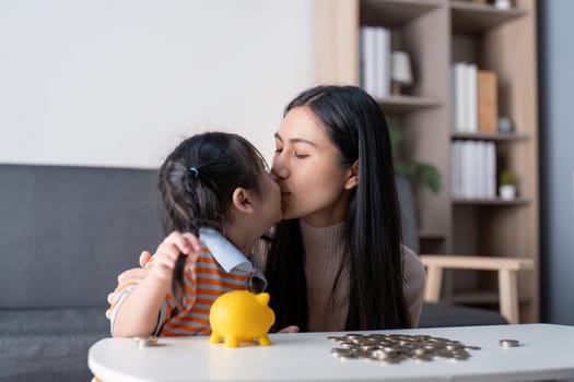 A diverse mother and her daughter share a sweet kiss as they engage in saving money together with a yellow piggy bank and coins, enhancing their family bond.