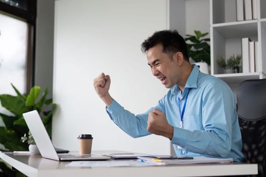 A joyful Asian businessman celebrates a success moment at his desk, raising his fists in excitement while working on a laptop in a contemporary office.
