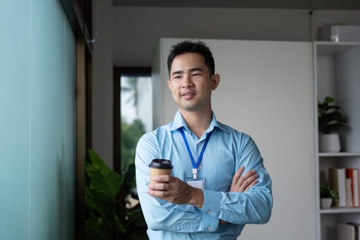 A confident Asian businessman stands with arms crossed, holding a coffee cup by a window in a contemporary office environment.