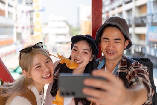 A group of young Asian friends smiling for a selfie with street food in hand, capturing happy moments in a bustling market atmosphere.