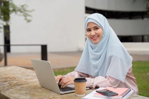 A joyful Muslim businesswoman in a hijab, focused on her laptop while enjoying a coffee, showcasing a blend of productivity and relaxation in an outdoor setting.