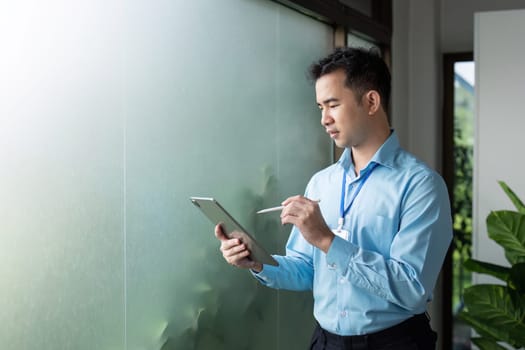 A focused Asian businessman is analyzing data on a tablet, standing by a frosted glass window in a contemporary office setting.