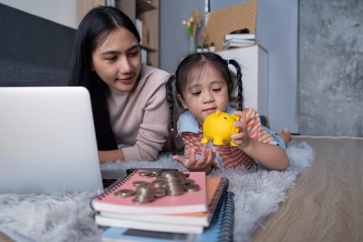 A diverse mother and her daughter examine a yellow piggy bank and coins together, enhancing their understanding of saving money while using a laptop at home.