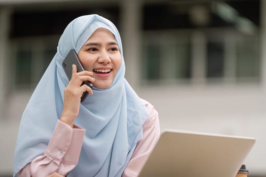 A joyful Muslim businesswoman engaged in a phone conversation, showcasing professionalism and modern work culture in an outdoor setting.