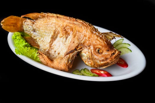 Fish grilled in batter with vegetables and greens on white plate isolated on black background side view