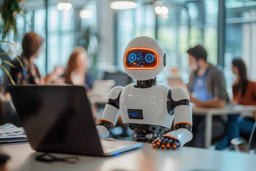 A robot is sitting in front of a laptop computer in a room with people. The robot is wearing a white suit and has a blue face. The people in the room are working on their laptops