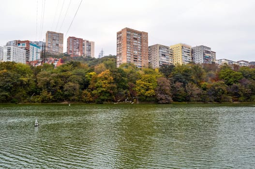 Dull and bleak view of old multi-story houses, glum sky, autumn forest and the lake