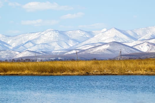 Stunning landscape of snowy mountains, dry grass against of lake, blue sky with clouds on a spring day. Russia, Far East