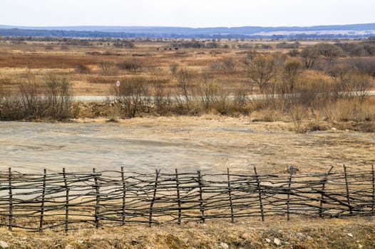Dull, depressing, gloomy landsape of field of dry grass, horses grazing in the distance and old wooden fence. 
Road without cars and speed limit sign. Copy space, without people