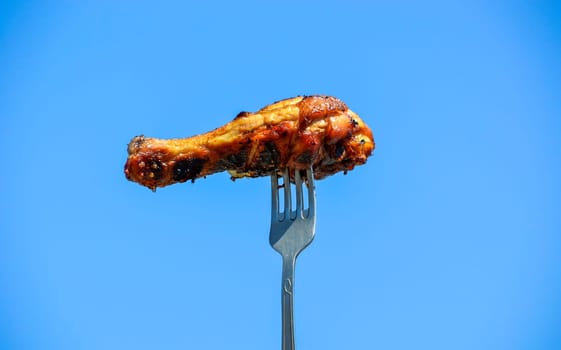 Grilled appetizing chicken leg on the fork against the blue sky. Outdoor barbecue
