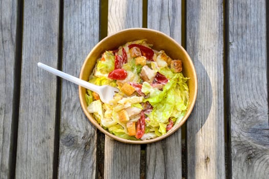 Paper bowl of fresh caesar salad with a fork on a wooden table. takeaway food