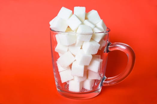 Transparent mug full of refined white sugar cubes on red background