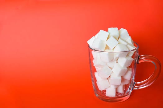 Transparent mug full of refined white sugar cubes on red background