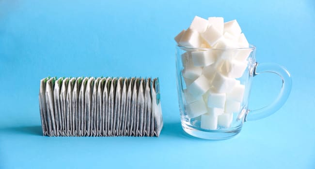 Many tea bags in a row next to a transparent glass mug full of white refined sugar on blue background