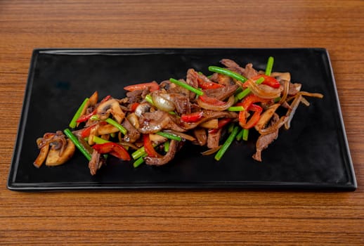 Fried pork and beef with garlic arrows, mushrooms and vegetables on a black plate on a wooden table.