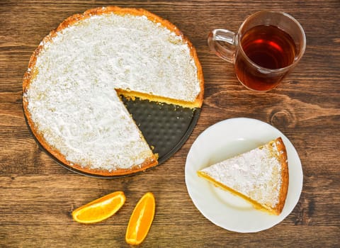 Piece of homemade pie with lemon and orange filling, covered with powdered sugar on a wooden table near orange fruit and cup of tea