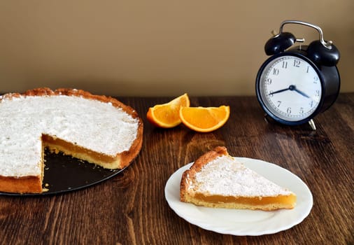 Piece of homemade pie with lemon and orange filling, covered with powdered sugar on a wooden table near orange fruit and alarm clock