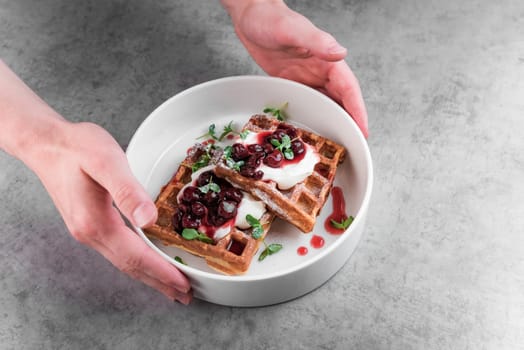 Waiter serving white plate with sweet belgian waffles with cream and cherry jam on a gray table in a cafe