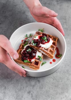 Waiter serving white plate with sweet belgian waffles with cream and cherry jam on a gray table in a cafe