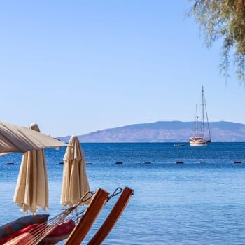 Beach umbrellas and sun loungers on the shore of a calm blue sea bay with a sailboat. Summer vacation and recreation area concept