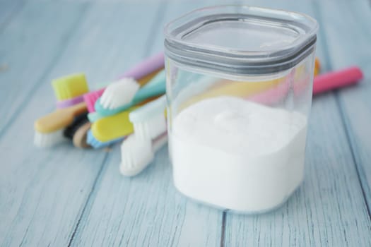 A wooden bowl of baking soda, a bamboo toothbrush, and a glass jar are displayed on a blue checkered fabric