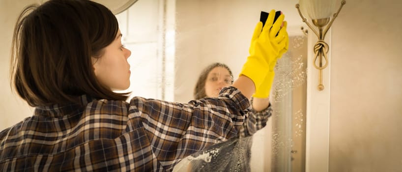 A young girl is cleaning the bathroom, applying detergent with a spray and washing the mirror with a sponge in yellow gloves on her hands. Smiling woman taking care of the cleanliness of her home.