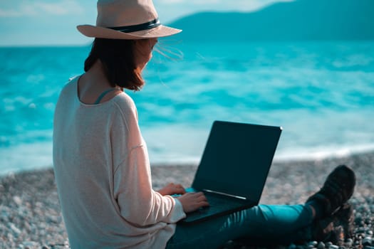 A young girl in a light hat and casual sweater lies on the beach by the sea with a laptop on a sunny day, works, studies, buys tickets during trip, a woman rests on vacation and types on the keyboard.