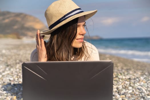 A young girl in a light hat and casual sweater lies on the beach by the sea with a laptop on a sunny day, works, studies, buys tickets during trip, a woman rests on vacation and types on the keyboard.