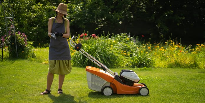 A young girl in a straw hat is mowing a lawn in the backyard with an orange lawn mower. A woman gardener is trimming grass with the grass cutter. A lawnmower is cutting a lawn on a summer sunny day.