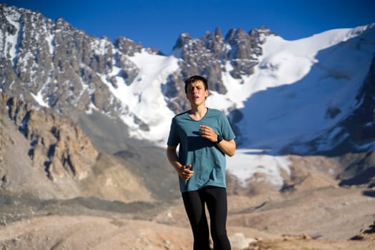 A young man runs along a trail in the snow-capped mountains, doing outdoor workout. The runner warms up, prepares for the race, leads a healthy active lifestyle.