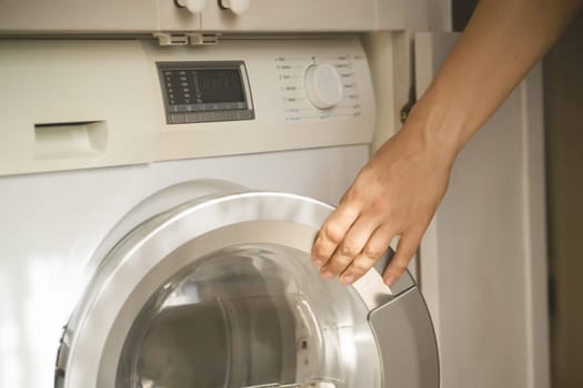 A male hand opens the door of a new washing machine in his modern, bright bathroom, close-up. The man washes clothes in the laundry.